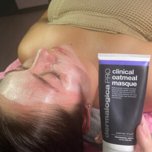 A woman laying on a table, receiving a facial treatment. There is a black tube next to her labeled "clinical oatmeal mask."