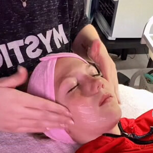 A child in a red robe with a pink band protecting their hair, laying back and receiving a face massage from an adult woman.